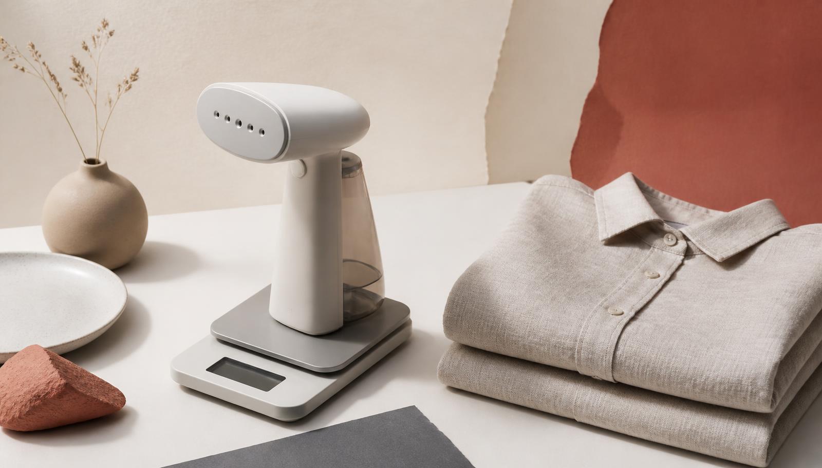 A lightweight handheld steamer on a kitchen scale beside folded linen shirts